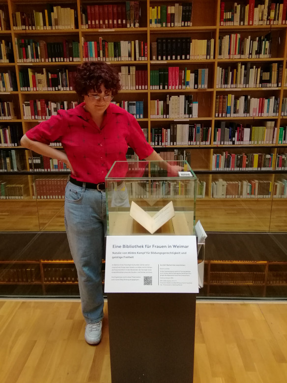 Emily Stopp neben der Vitrinenpräsentation "Eine Bibliothek für Frauen in Weimar" im Bücherkubus des Studienzentrums der Herzogin Anna Amalia Bibliothek